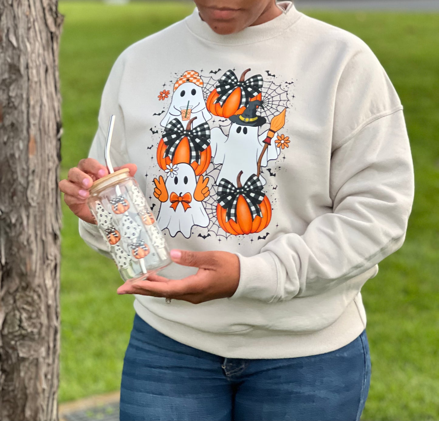 A woman holding a halloween glass can and wearing a matching halloween, crewneck sweatshirt with adorable ghosts and pumpkins with gingham bows.