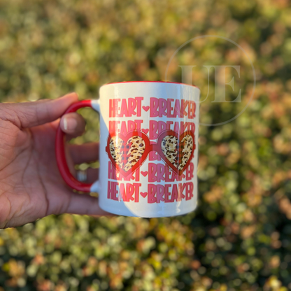 Image includes an 11oz white ceramic mug with red handle and interior with the text HeartBreaker on the front and an image of 2 hearts with cheetah prints inside.