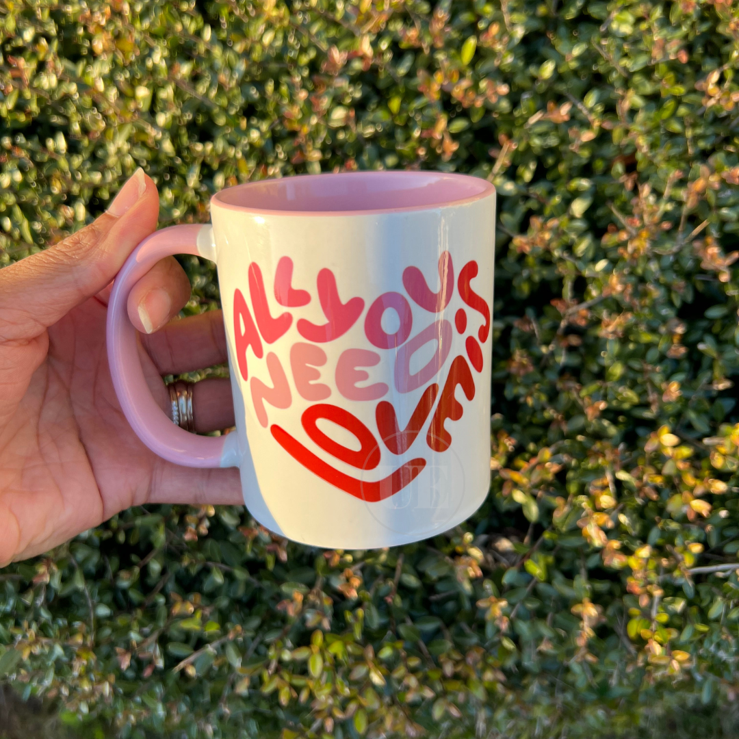 This is an image of an 11oz white ceramic mug with a pink handle and interior.  The front of the mug includes a multi-pink shaded text that says All You Need Is Love.